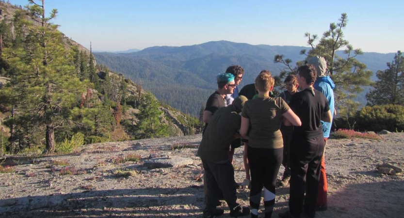 A group of people stand in a circle holding hands on top of a rocky overlook. There are tree covered mountains below. 